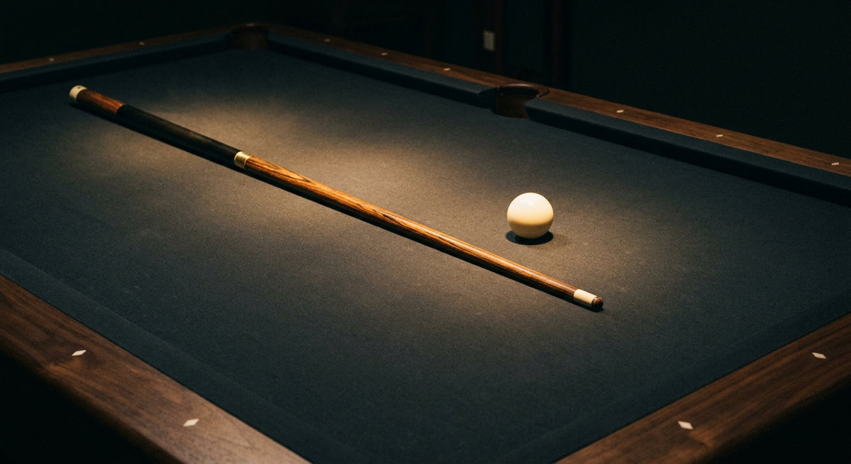 A polished pool cue resting diagonally across a dark billiard table with a single white cue ball under warm focused lighting.